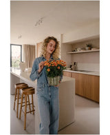 Eine Frau mit lockigen blonden Haaren, die ein Jeanshemd und Jeans trägt, steht in einer modernen Küche und hält eine künstliche Gänseblümchen-UV-Pflanze von 31 cm mit leuchtend orangefarbenen Blüten. Neben ihr stehen zwei Holzhocker auf der Kücheninsel.