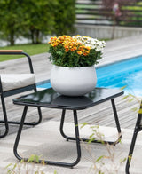 Ein schwarzer Terrassentisch mit einer künstlichen Gänseblümchen-UV-Pflanze (31 cm) – ein runder weißer Topf mit leuchtend orangefarbenen und weißen Gänseblümchen – steht neben einem Swimmingpool, umgeben von Gartenstühlen und mit viel Grün im Hintergrund.