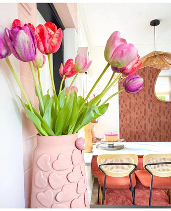 Eine rosa Vase mit geprägten Herzen hält die Kunstblume Französische Tulpe 60 cm rot auf einem Esstisch. Dahinter stehen Stühle mit Korbrücken, ein Kuchen auf dem Tisch und ein Spiegel an einer gemusterten braunen Wand unter einer geflochtenen Hängelampe.