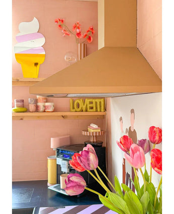 In einer gemütlichen rosa Küche steht im Vordergrund eine künstliche französische Tulpe (60 cm) in Rot, auf einem Regal pastellfarbene Tassen, ein LOVETTI-Teller, ein Wandgemälde mit zwei Personen und ein skurriler Eistütenspiegel an der Wand.