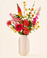 In einer weißen Keramikvase befindet sich der Strauß „Summer Blush“ aus künstlichen Seidenblumen, bestehend aus rosa, gelben und orangefarbenen Blüten mit grünen Blättern, vor einem schlichten hellen Hintergrund.