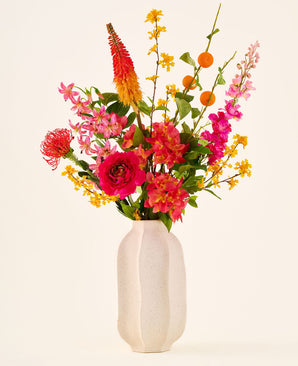 In einer weißen Keramikvase befindet sich der Strauß „Summer Blush“ aus künstlichen Seidenblumen, bestehend aus rosa, gelben und orangefarbenen Blüten mit grünen Blättern, vor einem schlichten hellen Hintergrund.