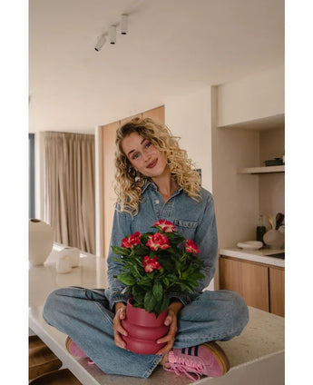 Eine Frau mit lockigem blondem Haar, trägt einen Jeansoverall und rosa Turnschuhe, sitzt mit gekreuzten Beinen auf einer Küchentheke und hält lächelnd die künstliche Gardenie UV 39 cm in Fuchsia in der Hand.