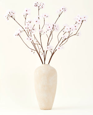 Der Seiden-Kunstblumenstrauß Kirschblüten hellrosa 114 cm steht in einer hohen beigen Keramikvase mit langen, dünnen Zweigen und zarten hellrosa Blüten und setzt einen ruhigen, künstlichen Blumenakzent vor einem schlichten Hintergrund.