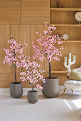 Drei Topfpflanzen vom Typ Kunstpflanze Bougainvillea, 120 cm groß, rosafarben und mit rosa Blüten, stehen auf einem weißen Boden in einem modernen Raum mit Holzvertäfelung, Regalen und einem weißen Stuhl.