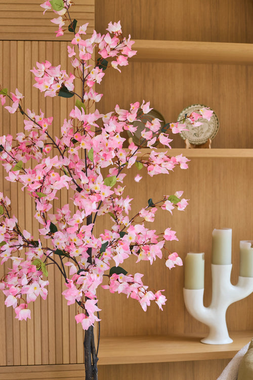 Eine künstliche Bougainvillea-Pflanze von 180 cm, rosafarben mit rosa Blüten, steht vor Holzregalen, auf denen ein dekorativer Teller und ein weißer Kerzenständer mit drei grünen Kerzen stehen.