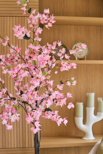 Vor den Holzregalen, auf denen ein Teller und ein weißer Kerzenständer mit drei grünen Kerzen ausgestellt sind, steht eine 120 cm große künstliche Bougainvillea-Pflanze in Rosa.