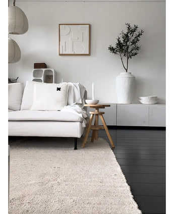 Ein minimalistisches Wohnzimmer mit einem hellen Sofa, einem neutralen Teppich, einem weißen Sideboard, einem künstlichen Olivenbaum 120 cm als Dekoelement, abstrakter Wandkunst und Holzhockern – alles in einer sanften mediterranen Atmosphäre mit neutralen Tönen.