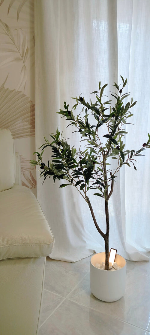 Ein künstlicher Olivenbaum 120 cm mit grünen Blättern steht auf einem Fliesenboden neben einem cremefarbenen Sofa und verleiht dem Raum eine mediterrane Atmosphäre, vor weißen Vorhängen und einer Wand mit dezentem Muster.