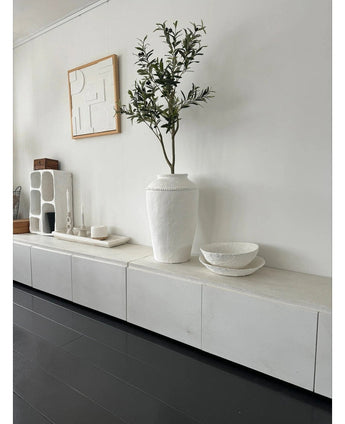 Ein minimalistischer Raum mit einem weißen Sideboard, dem künstlichen Olivenbaum 120 cm als Dekoelement, dekorativen weißen Schalen, einer Kerze und abstrakter Wandkunst. Dunkle Holzböden und moderne Akzente schaffen eine ruhige mediterrane Atmosphäre.
