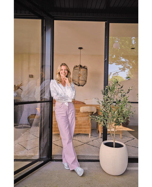 Eine Frau in weißer Bluse und lavendelfarbener Hose lächelt neben einer großen Glastür. Der künstliche Olivenbaum 120 cm bringt mit einem Korbsofa und hellem Dekor im Hintergrund eine mediterrane Atmosphäre in das gemütliche Wohnzimmer.