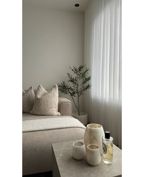 Ein minimalistisches Wohnzimmer verfügt über ein beiges Sofa, strukturierte Kissen, einen künstlichen Olivenbaum 120 cm als Dekoration, transparente weiße Vorhänge und einen Marmortisch mit drei cremefarbenen Vasen und Parfüm. Natürliches Licht erhellt den ruhigen Raum.