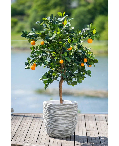 Ein 120 cm großer künstlicher Orangenbaum mit reifen Früchten steht in einem weiß geprägten Topf auf einer Holzterrasse mit Blick auf ein dunstiges Wasserspiel und eine grüne Landschaft draußen.
