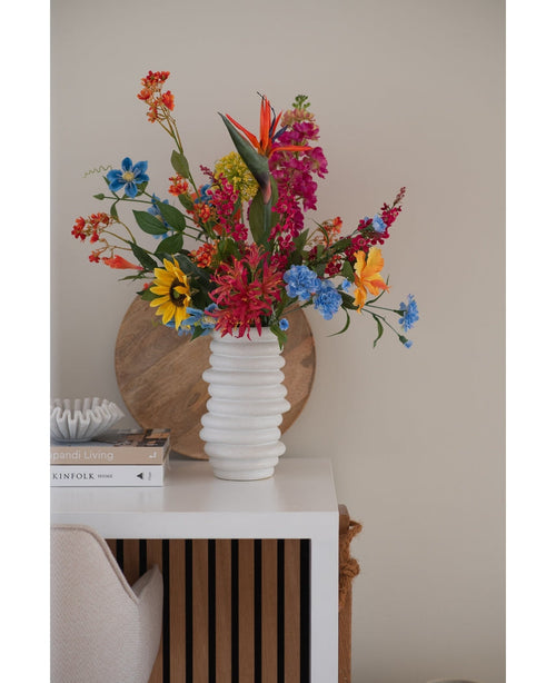 Auf einem weißen Tisch neben gestapelten Büchern und Deko steht in einer weißen, strukturierten Vase ein künstlicher Blumenstrauß aus Katzenminze in Rosa (75 cm), im Hintergrund lehnt ein rundes Holztablett an der beigen Wand.