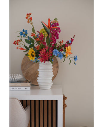 Auf einem weißen Tisch neben gestapelten Büchern und Deko steht in einer weißen, strukturierten Vase ein künstlicher Blumenstrauß aus Katzenminze in Rosa (75 cm), im Hintergrund lehnt ein rundes Holztablett an der beigen Wand.