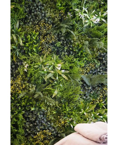 Eine üppige, strukturierte Kulisse wird durch die vertikale UV-Gartenwand Kunsthaag Freshness 100x100 cm geschaffen, die dicht mit grünem Blattwerk bedeckt ist. Unten rechts sind zwei gefaltete beige Handtücher zu sehen.