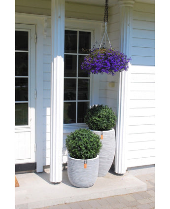 Auf der weißen Veranda eines Hauses befinden sich zwei UV-Topiary-Pflanzgefäße im römischen Stil (65 cm) mit künstlichem Buchsbaum auf Stiel und grünen Sträuchern, darüber ein Hängekorb mit violetten Blumen, weiße Säulen und eine Glastür auf der linken Seite.