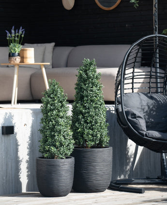 Zwei künstliche Buchsbaumpyramiden UV 120 cm werden auf einer Holzterrasse vor einem Sitzbereich im Freien mit einem hellen Sofa, einem runden Tisch mit Lavendel und einem schwarzen Hängesessel platziert.