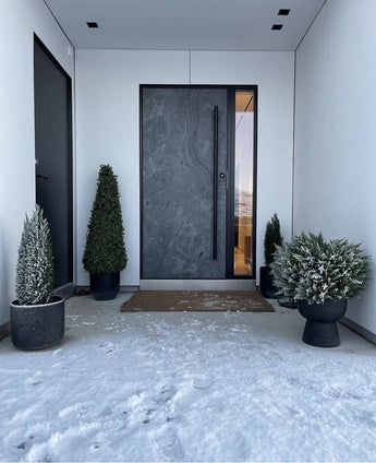 Eine moderne dunkle Haustür wird von weißen Wänden eingerahmt und flankiert von einer künstlichen Buchsbaumpyramide UV 90 cm, die UV-Schutz bietet. Leichter Schnee bedeckt den Boden, während diese stilvollen Gartenpflanzen einen aufgeräumten, minimalistischen Eingangsbereich schaffen.