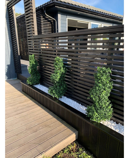 Drei künstliche Buchsbaumspiralen im römischen UV-Stil (90 cm) stehen in einem schmalen Bett aus weißen Kieselsteinen vor einem modernen schwarzen Lattenzaun neben einer Holzterrasse und einem modernen Haus.
