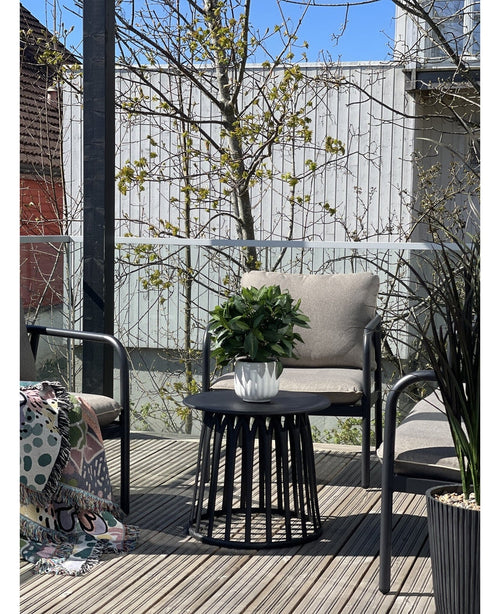 Eine moderne Terrasse mit zwei Stühlen mit Kissen, einem runden schwarzen Tisch mit der künstlichen Lorbeerpflanze UV d33 cm als Mittelpunkt und einer gemusterten Decke auf einem Stuhl. Im Hintergrund erscheinen unter klarem Himmel Bäume und ein Holzzaun.