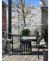 Eine moderne Terrasse mit zwei Stühlen mit Kissen, einem runden schwarzen Tisch mit der künstlichen Lorbeerpflanze UV d33 cm als Mittelpunkt und einer gemusterten Decke auf einem Stuhl. Im Hintergrund erscheinen unter klarem Himmel Bäume und ein Holzzaun.