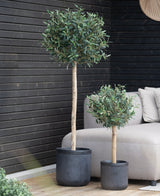 Zwei künstliche Olivenbäume UV 85 cm in einem Topf mit rundem, blattgrünem Blattwerk stehen auf einer Holzterrasse neben einem beigen Outdoor-Sofa und einer Wand mit schwarzen Paneelen. Der größere Baum steht in einem hohen Topf, der kleinere in einem kürzeren Topf.