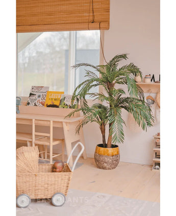 In einem gemütlichen, hellen Raum werden die Kunstpalme Cycas Classic 135 cm in einem stilvollen Pflanzgefäß, einem Spielzeugwagen aus Korbgeflecht, Holzmöbeln und Kinderbüchern neben einem Fenster mit einem Bambusrollo platziert.