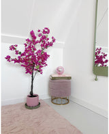 Eine moderne, minimalistische Raumecke mit einer künstlichen Bougainvillea (125 cm) in Lila in einem Topf, einem runden rosa Fußhocker mit goldenen Beinen, gestapelten Büchern, einer rosa Kristallkugel, einem Wandspiegel und einem zartrosa Teppich auf einem hellen Boden.