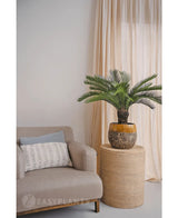 Eine künstliche Cycas-Palme 80 cm steht auf einem runden Korbtisch neben einem beigen Sessel mit weiß gemustertem Kissen, vor hellbeigen Vorhängen und einer neutralen Wand.