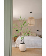Ein minimalistisches Schlafzimmer mit beigen Wänden, einem weißen Bett, einem natürlich gewebten Teppich und der künstlichen Pflanze Enkianthus 210 cm – einer großen Topfpflanze mit grünen Blättern – im Vordergrund. Von der Decke hängt eine gewebte Hängelampe.