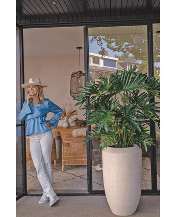 Eine Frau in blauer Bluse, weißer Hose und Hut lacht über eine Glastür neben der künstlichen Pflanze Philodendron 125 cm in einem beigen Topf. Korbstühle und Dekor sind im ganzen Raum sichtbar.