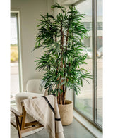 Die künstliche Marienpalme 210 cm mit ihren langen, schmalen grünen Blättern wird in der Nähe großer Fenster in einem hellen Raum platziert. Ein weißer Sessel und ein mit einer hellen Decke drapierter Holzstuhl daneben sorgen für eine einladende Atmosphäre.