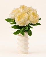In einer gewellten, strukturierten weißen Vase befindet sich der Seiden-Kunstblumenstrauß Peony White – große cremefarbene künstliche Pfingstrosenblüten mit grünen Blättern vor einem schlichten hellen Hintergrund.
