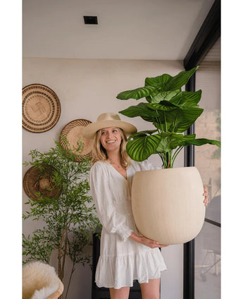 Eine Frau in einem weißen Kleid und Hut lächelt, während sie die künstliche Calathea UV-Pflanze (72 cm) in den Innenbereich hält, mit geflochtenen Körben und einer weiteren Pflanze im Hintergrund.