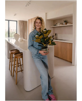 Eine lächelnde Person in Jeanskleidung steht in einer modernen Küche und hält eine künstliche Magnolie in einem 46-cm-Topf, eine pflegeleichte Pflanze. Die Küche eignet sich perfekt für die Inneneinrichtung und verfügt über Holzschränke, eine weiße Kücheninsel und zwei Barhocker.