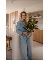 Eine Frau mit lockigen blonden Haaren und Jeanskleidung steht lächelnd in einer modernen Küche mit der künstlichen Magnolienpflanze in einem 46-cm-Topf in ihren Händen, umgeben von Holzschränken und Regalen – eine ideale Wahl für alle, die nach pflegeleichten Pflanzen suchen, um ihr stilvolles Interieur aufzuwerten.