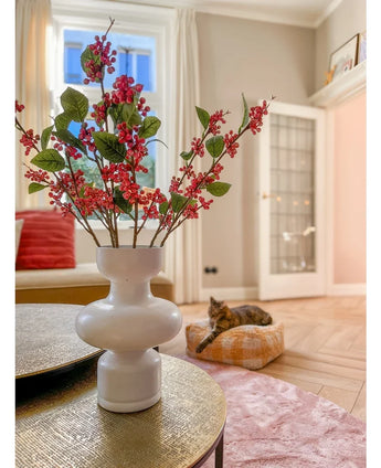 Auf einem Tisch steht eine weiße Vase mit rosa Kunsttak-Beeren (64 cm) und grünen Blättern. Im Hintergrund liegt eine Katze auf einem rosa Kissen in einem hellen Wohnzimmer mit beigen Wänden und einer Glastür.