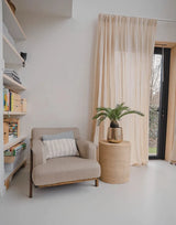 Eine gemütliche Leseecke mit einem beigen Sessel, einem runden Beistelltisch aus Korbgeflecht mit der künstlichen Baby-Cycas-Palme 60 cm darauf, Bücherregalen und Gardinen, die über einer Glastür hängen, die zum Garten führt.