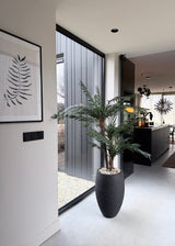 Ein modernes, minimalistisches Interieur mit einer künstlichen Cycas-Klassikpalme 135 cm in einem stilvollen dekorativen Topf in der Nähe eines raumhohen Fensters, einem Farnkunstwerk an der Wand und einer eleganten schwarzen Küche mit Hängelampen im Hintergrund.