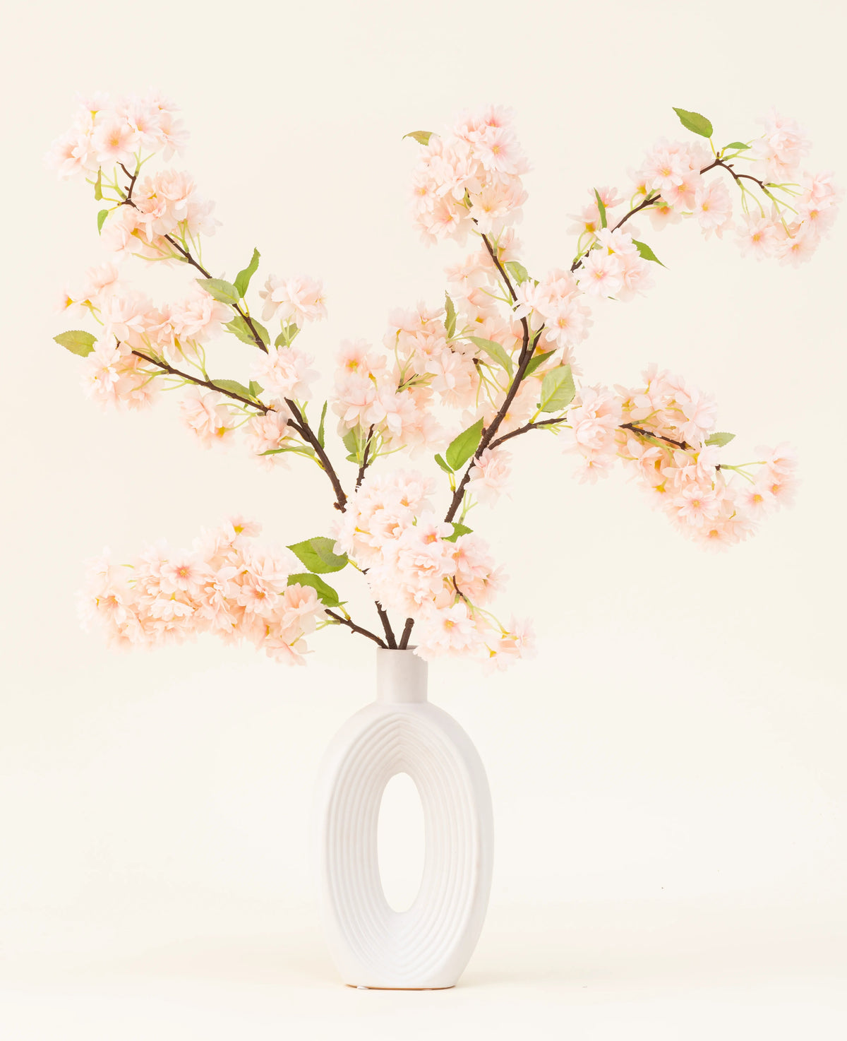 Eine weiße Keramikvase mit einer breiten ovalen Öffnung zeigt mehrere grünblättrige künstliche Seidenblumenstrauß hellrosa Kirschblütenzweige vor einem einfachen hellen Hintergrund.