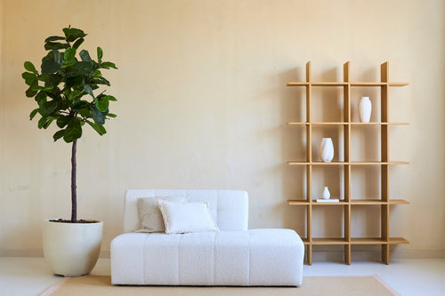 Minimalistisches Wohnzimmer mit einem weißen Sofa, zwei Kissen und einer Kunstpflanze Ficus Tabakpflanze Deluxe 180 cm neben einem offenen Holzregal mit weißen Vasen, alles vor einer beigen Wand und auf einem hellen Teppich.