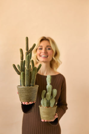 Eine lächelnde Frau in einem braunen Kleid hält zwei künstliche Kaktus-Cereus-Topfpflanzen (35 cm), eine große und eine kleine, vor einem schlichten beigen Hintergrund.