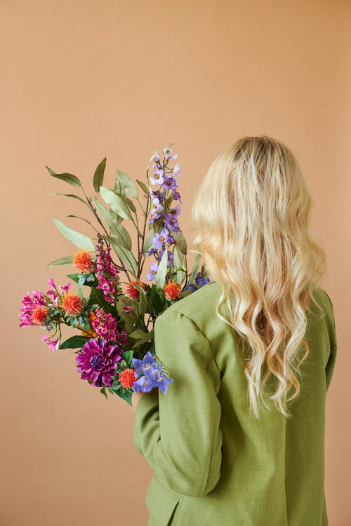Von hinten ist eine Person mit langen, welligen blonden Haaren und einer grünen Jacke zu sehen, die den farbenfrohen Kunstblumenstrauß aus Svenski-Seide vor einem schlichten beigen Hintergrund hält.