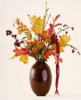 In einer braunen Vase befindet sich der Strauß „Rustic Glow“ aus künstlichen Seidenblumen mit gelben, orangefarbenen und roten Blättern, roten Beeren, orangefarbenen Blüten und langem, hängendem rotem Amaranthus vor einem neutralen Hintergrund.