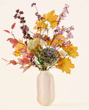 Der Strauß „Wilder Amethyst“ aus künstlichen Seidenblumen mit Herbstblättern, violetten und weißen Blüten und grünen Hortensien steht in einer weißen Keramikvase vor einem schlichten, hellen Hintergrund.