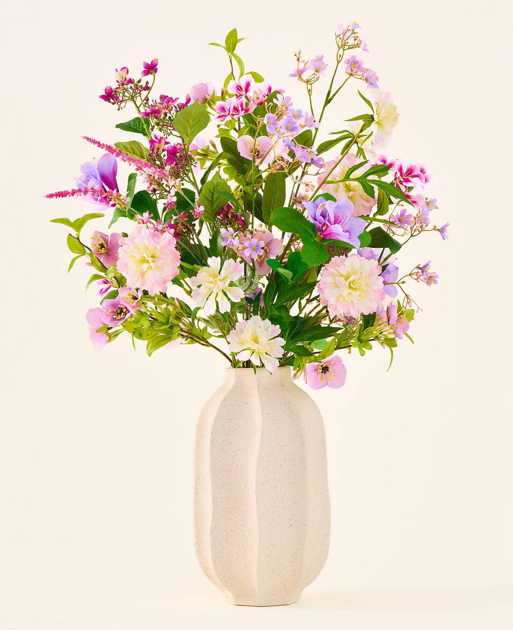 Eine weiße Keramikvase enthält einen farbenfrohen Strauß aus rosa, lila und weißen Blumen aus der Kollektion „Seiden-Kunstblumenstrauß Royal Bloom“, akzentuiert mit grünen Blättern und präsentiert vor einem schlichten, hellen Hintergrund.