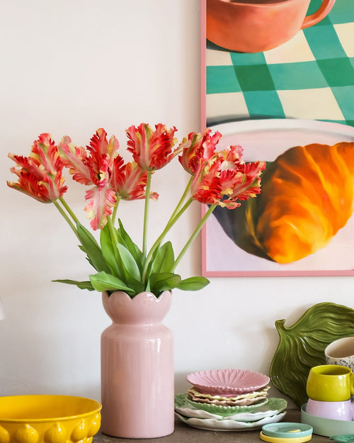 Auf einem Tisch mit buntem Geschirr steht eine rosa Vase mit der Kunstblume Französische Sittich-Tulpe 76 cm rot/gelb, während im Hintergrund teilweise ein gerahmtes Gemälde mit einem Croissant und einer Tasse zu sehen ist.