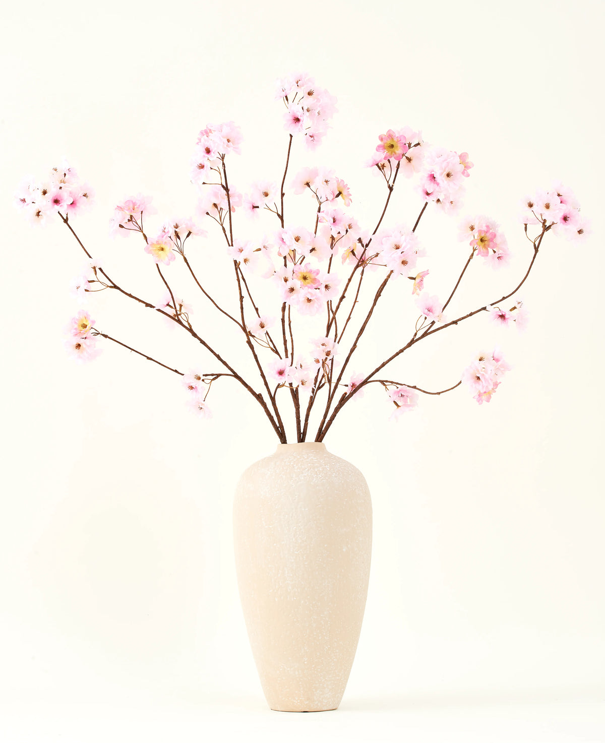 Der Seiden-Kunstblumenstrauß Kirschblütenrosa 114 cm hat lange, dünne Zweige mit Büscheln hellrosa Blüten in einer beigen Keramikvase, die einen auffälligen Kontrast zum schlichten weißen Hintergrund bilden.