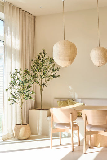 Ein heller, minimalistischer Essbereich ist mit zwei hellen Holzstühlen, einem kleinen Tisch, Papierlaternen und dem 180 cm großen künstlichen Olivenbaum ausgestattet, der einen Hauch von mediterranem Stil verleiht. Eingetopfte Zimmerbäume, durchscheinende beige Vorhänge und Sonnenlicht, das durch die großen Fenster fällt, runden diesen entspannenden Raum ab.